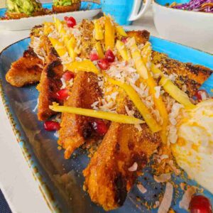 Golden Mango Coconut Captain Crunch French Toast Brioche topped with fresh pineapple mango cream cheese frosting, toasted coconut, and pomegranate, served with maple syrup.