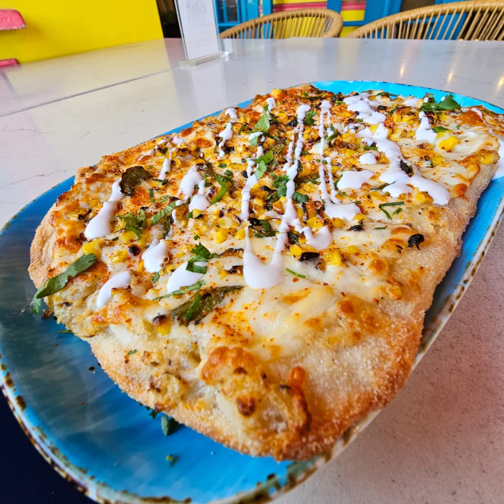 A whole rectangular elote flatbread pizza served on a textured blue plate at Nalu Vida Venice Beach, generously topped with roasted corn, crema, and fresh herbs.
